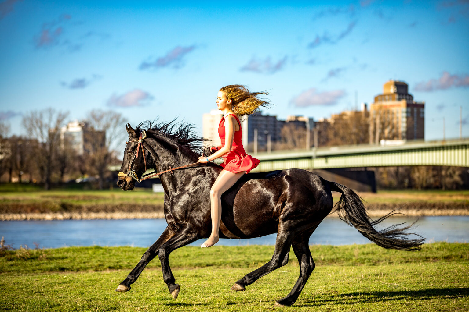 Teenager reitet im roten Kleid auf ihrem Pferd vor dem Rheinufer